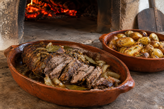 Javali assado fatiado em tabuleiro de barro, com batatas assadas junto a um forno de lenha