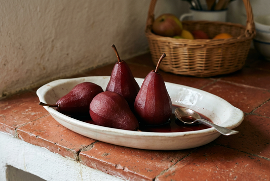 Quatro peras inteiras de cor rubi, com os pés, numa travessa branca com calda de vinho tinto, sobre uma prateleira rústica de terracota.